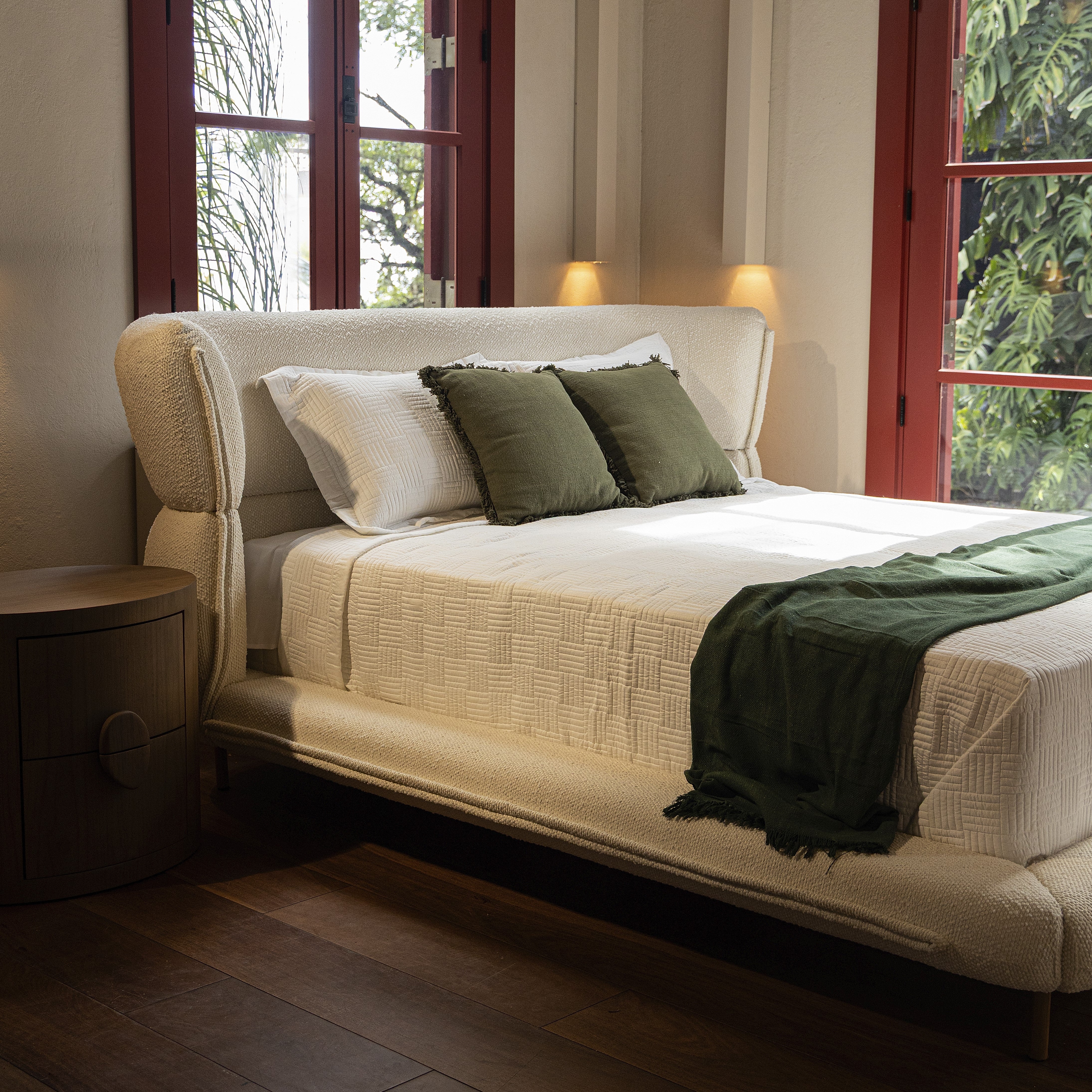 Beige bed with green pillows and blanket in a room with large windows.