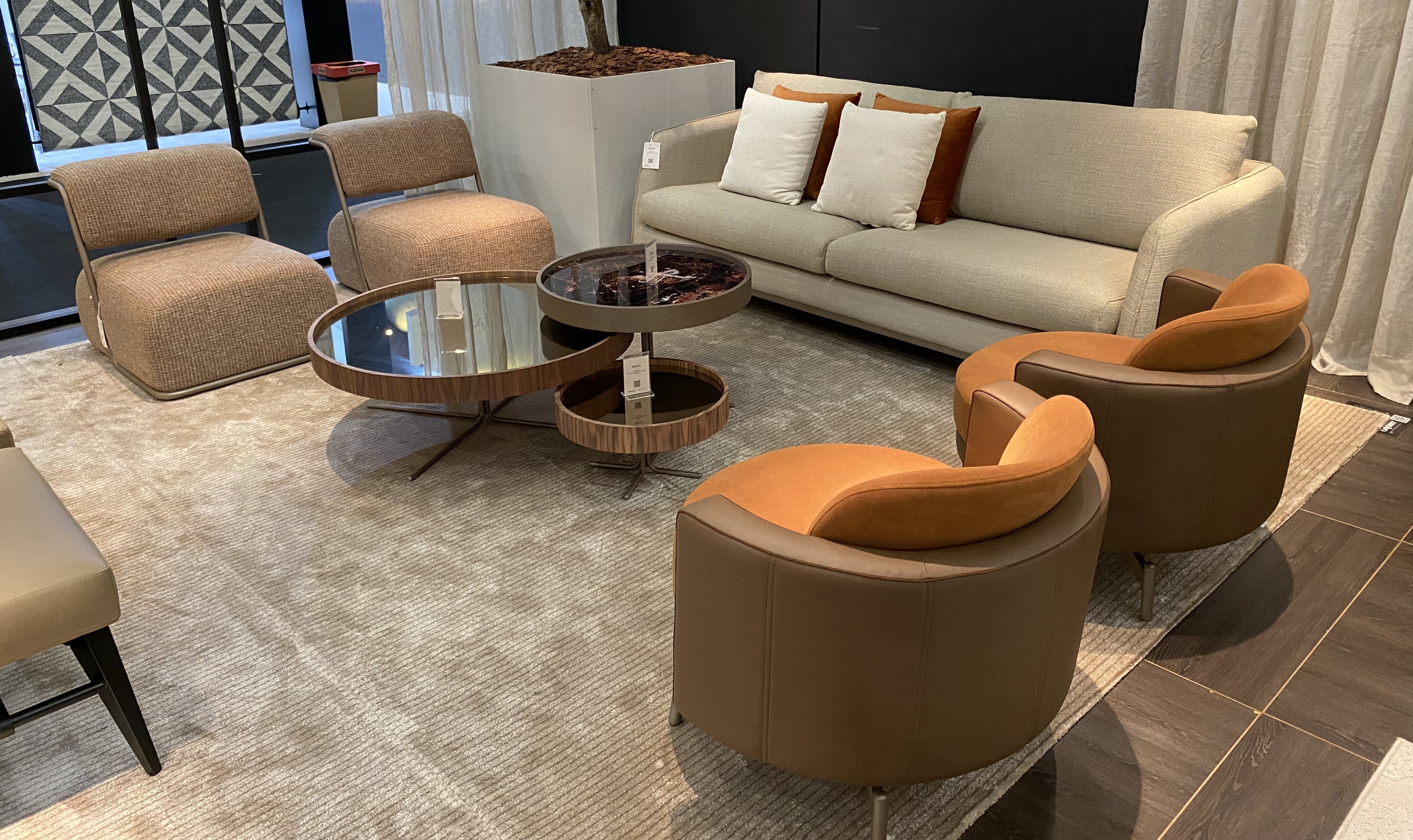 A modern living room furniture set featuring a cream-colored sofa with brown and white pillows, four upholstered armchairs in brown and tan leather, and two round nesting coffee tables on a large textured beige rug.