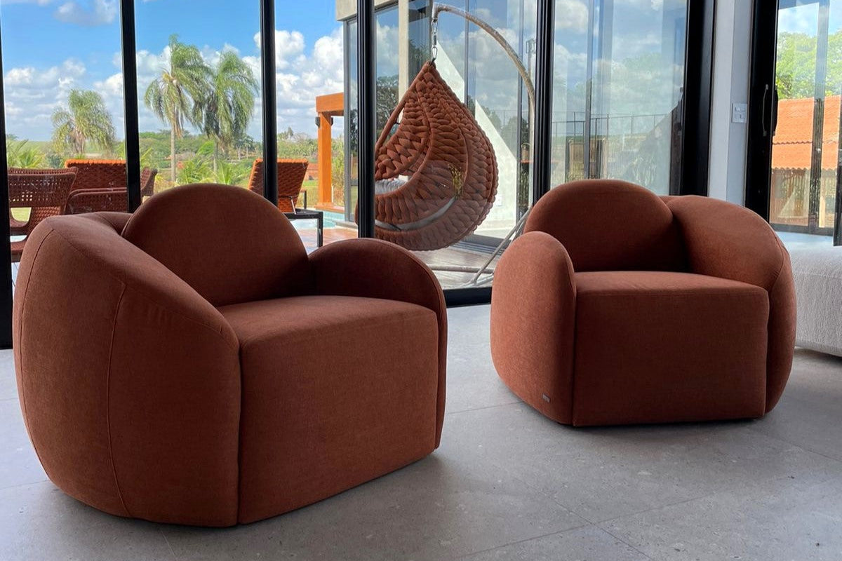 Two modern terracotta-brown armchairs with rounded, bubble-like forms placed in a bright interior with large floor-to-ceiling windows and a hanging wicker swing visible in the background.