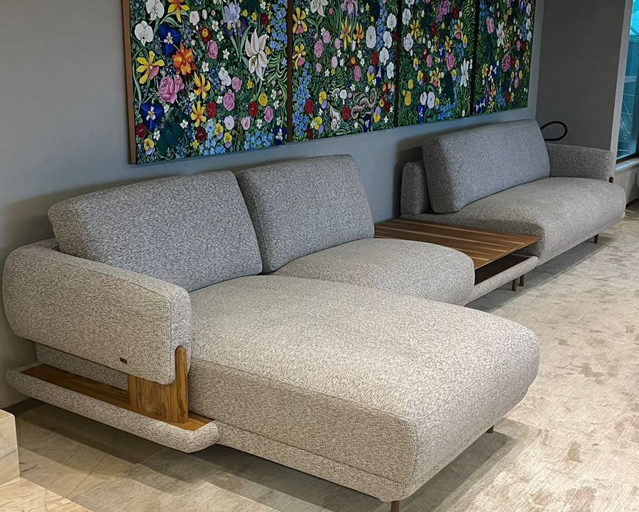 A modern light gray chaise lounge sofa with a wooden accent table, set in a living room environment with a large floral triptych wall art piece on the wall behind it.