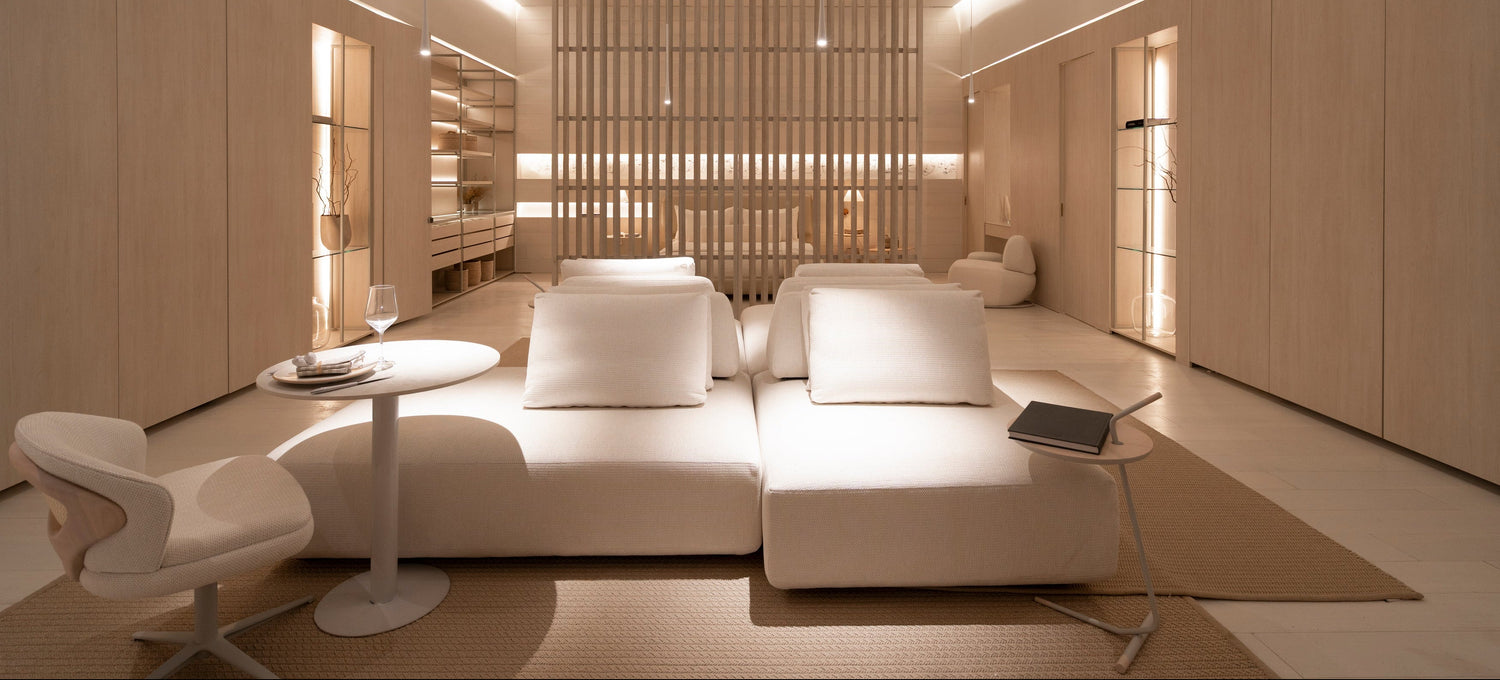 A modern minimalist interior featuring a large white modular sofa with white pillows, a small round white side table, a white sculptural chair, and light wood-paneled walls and ceiling with integrated linear lighting.