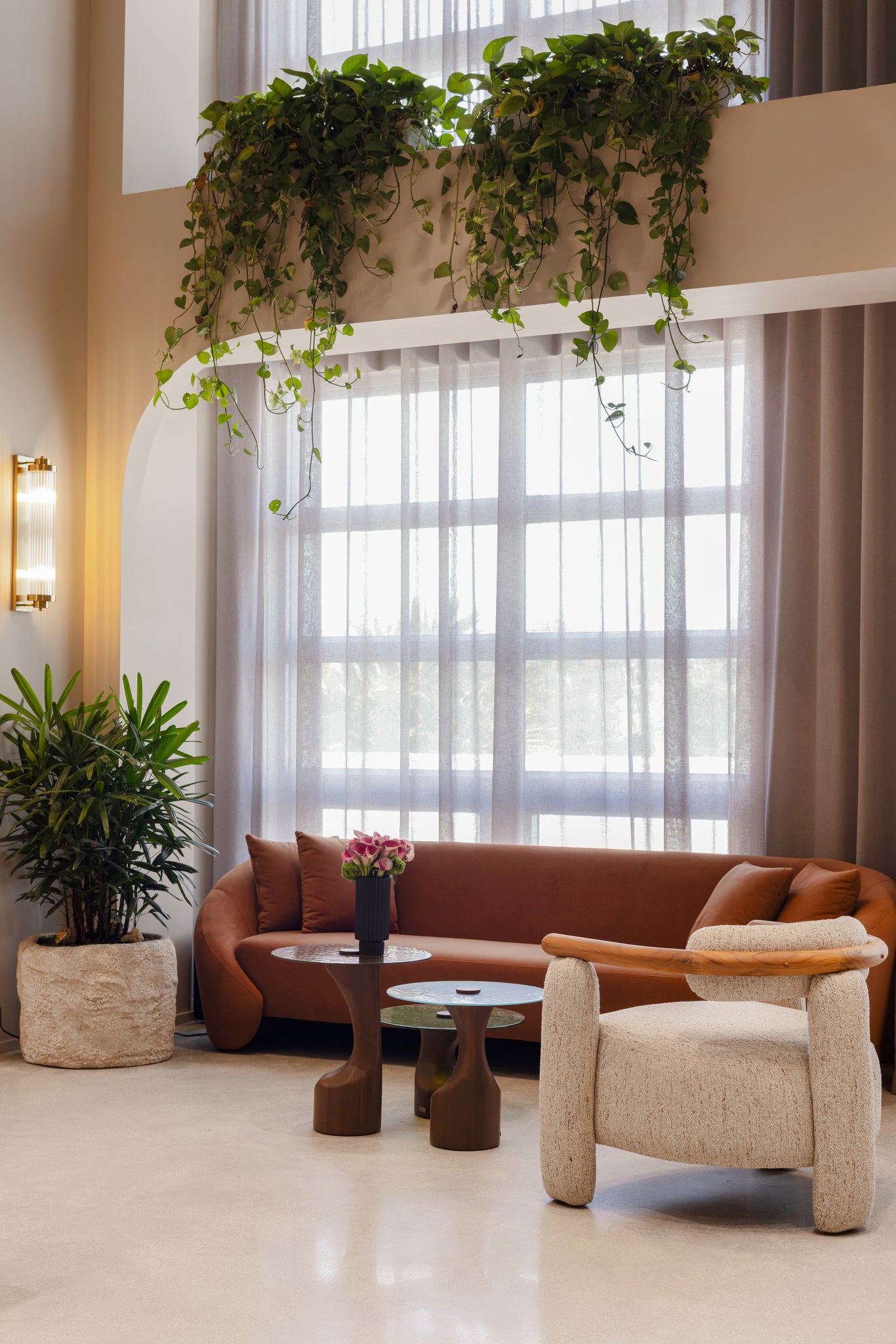 A modern living room interior featuring a rust-brown leather sofa, a cream boucle armchair, two sculptural dark wood coffee tables, a potted green plant, and cascading green trailing plants hanging from a high window.