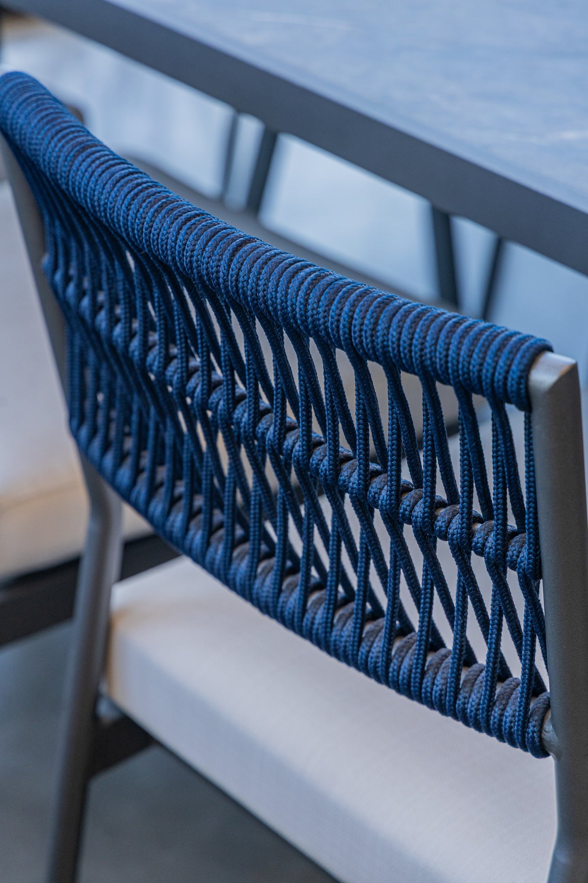 Close-up of a modern outdoor chair with a woven blue rope backrest and a light beige seat cushion, set next to a dark table.
