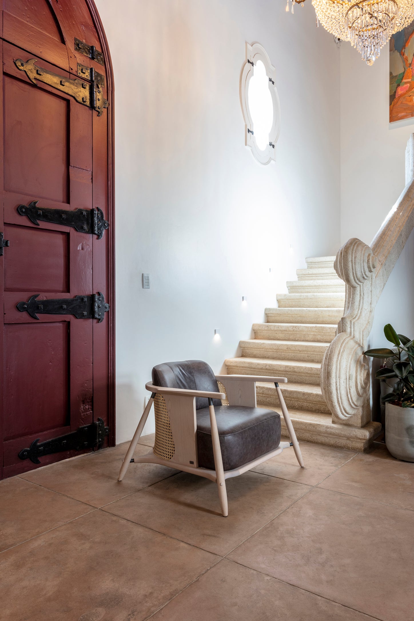 A modern mid-century style accent chair with a dark gray leather seat and light wood frame, positioned in a bright entryway next to a large dark wood door and a stone staircase.