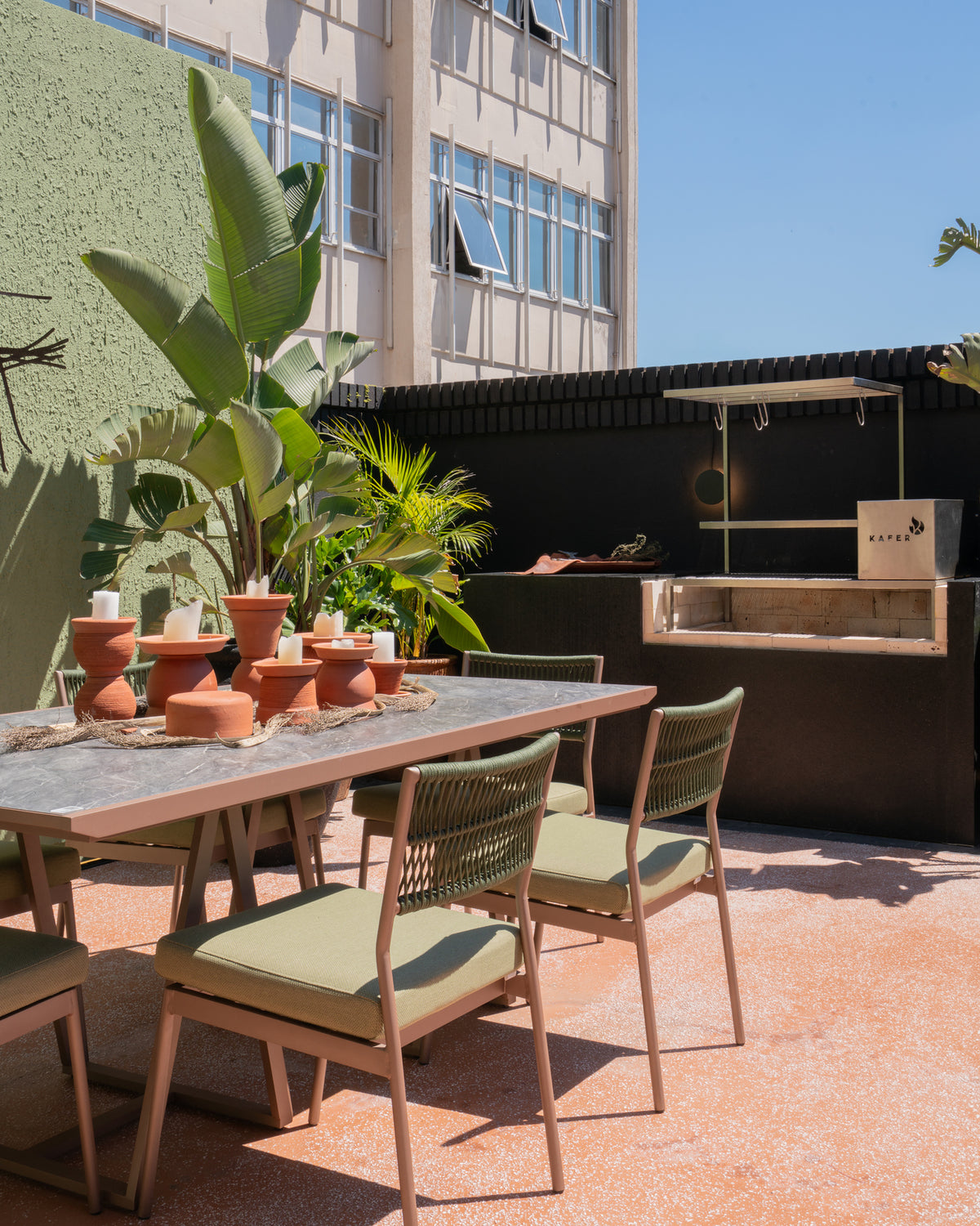 Outdoor dining area with a rectangular table and woven-back chairs under bright sunlight, set against a modern building wall with plants and a black counter.