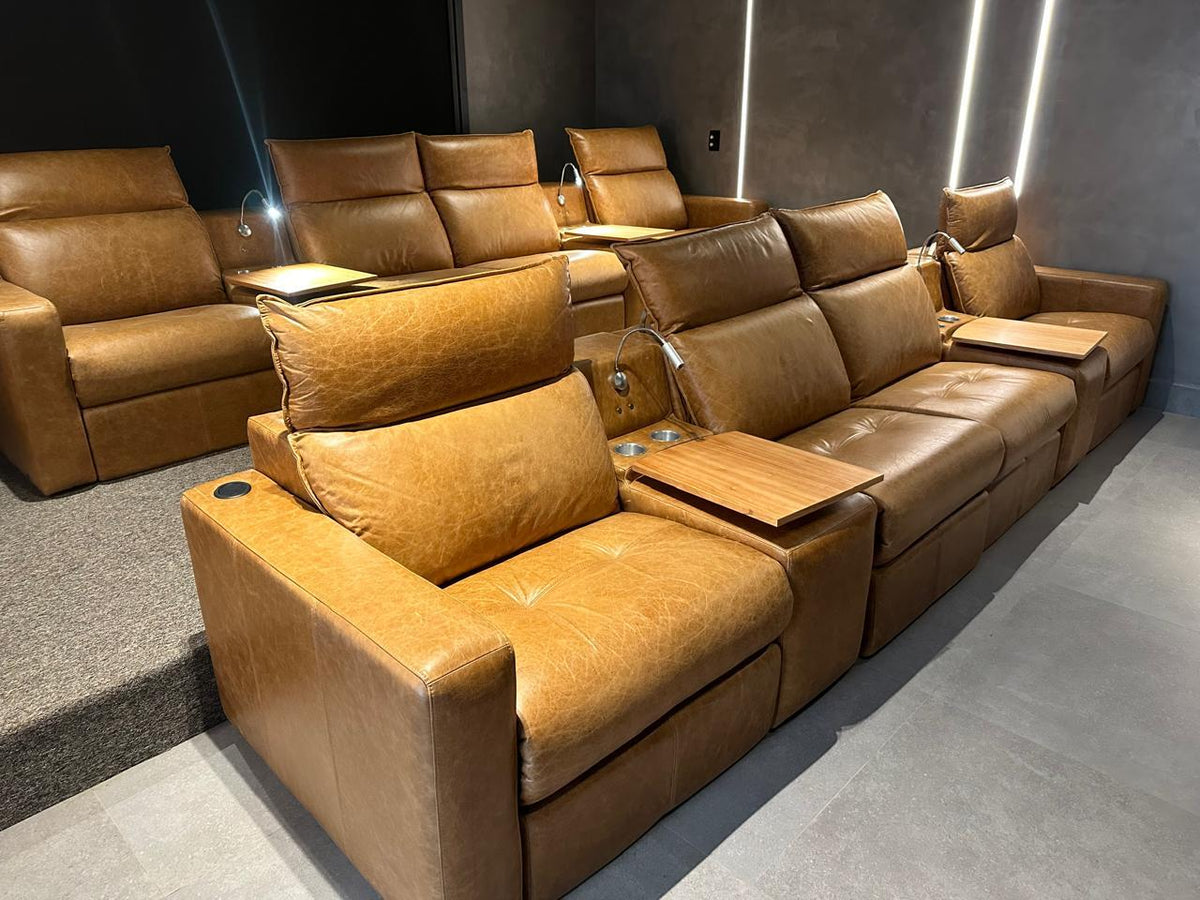 A large tan leather home theater seating system with multiple reclining seats, built-in floor speakers, and fold-out side tables, shown in a modern home theater setting with gray walls and ambient lighting.