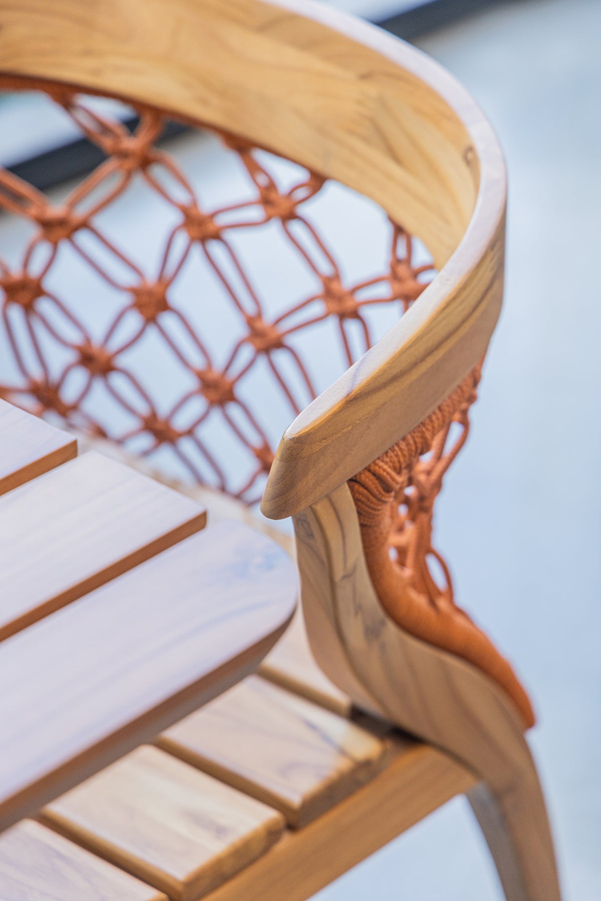 Close-up of a wooden outdoor chair with a curved armrest, light natural wood slats, and a terracotta-colored woven mesh backrest.