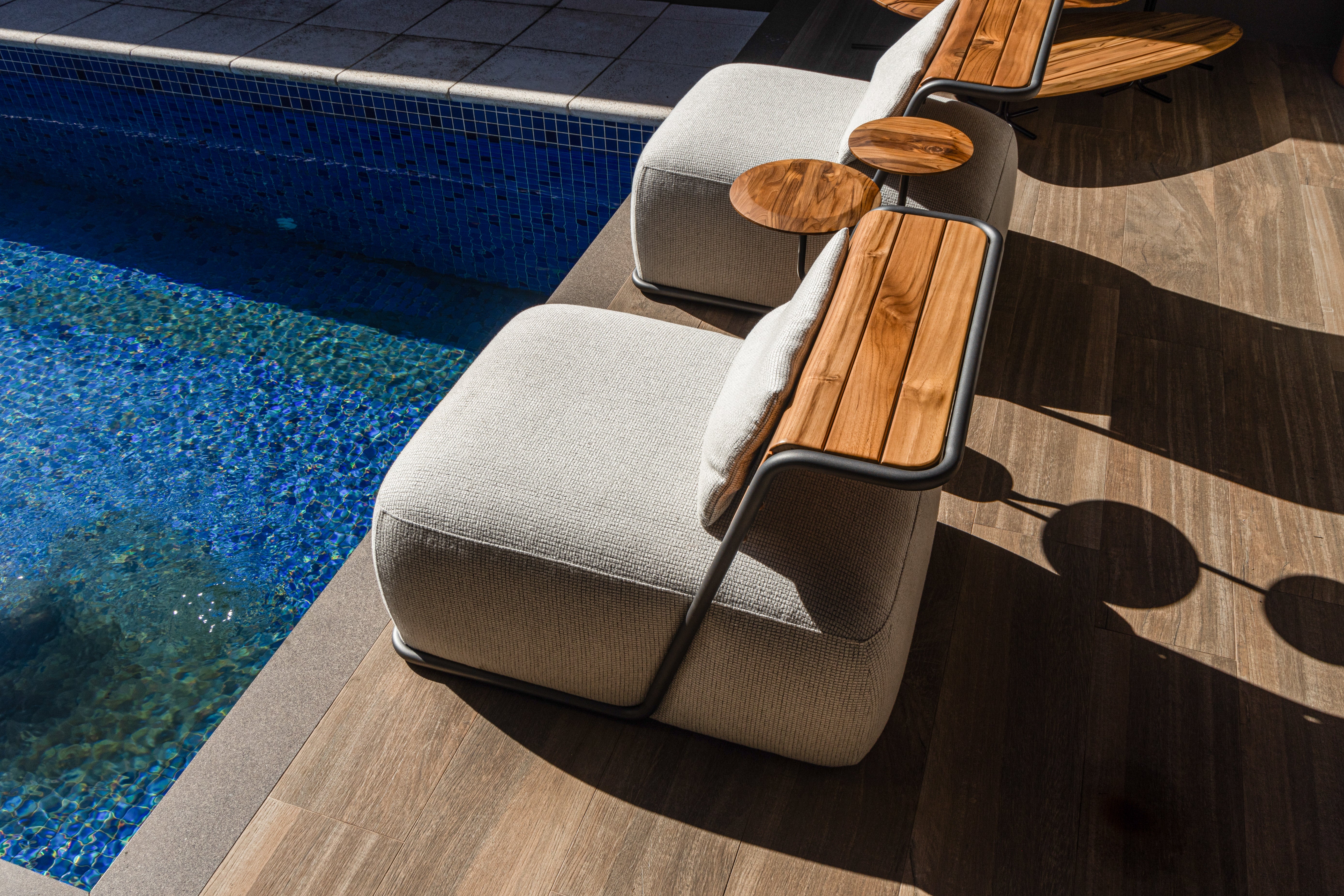 Modern outdoor lounge chair with a light beige textured fabric seat and backrest, a curved wooden armrest with a round wooden side table, and a white cushion, positioned next to a blue tiled swimming pool on a wooden deck.
