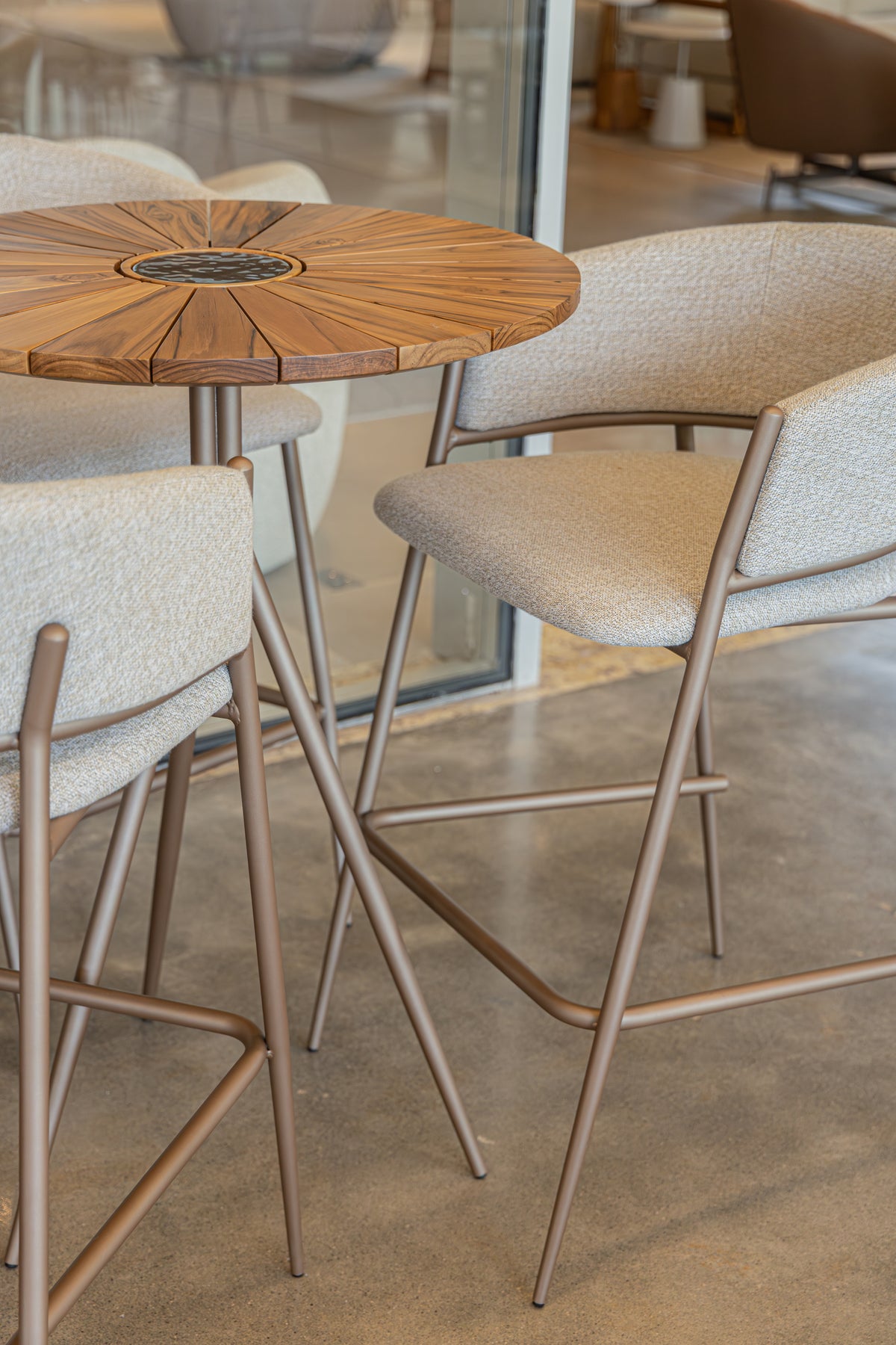 A modern round wooden bar-height table with a central inlay, paired with upholstered bar stools in a bright indoor setting with concrete floors.