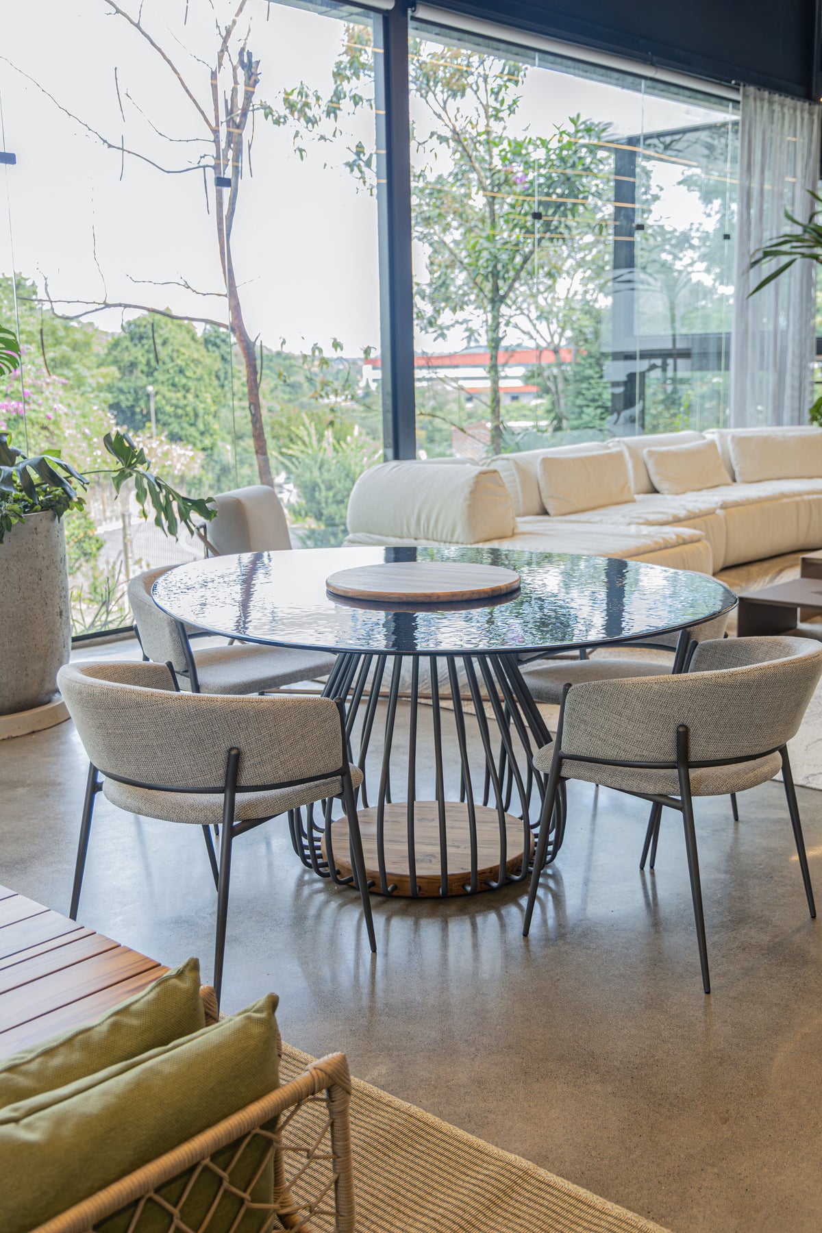 A modern dining set featuring a round glass-top table with a black wire base and four upholstered dining chairs, set in a contemporary living space with large windows overlooking greenery.