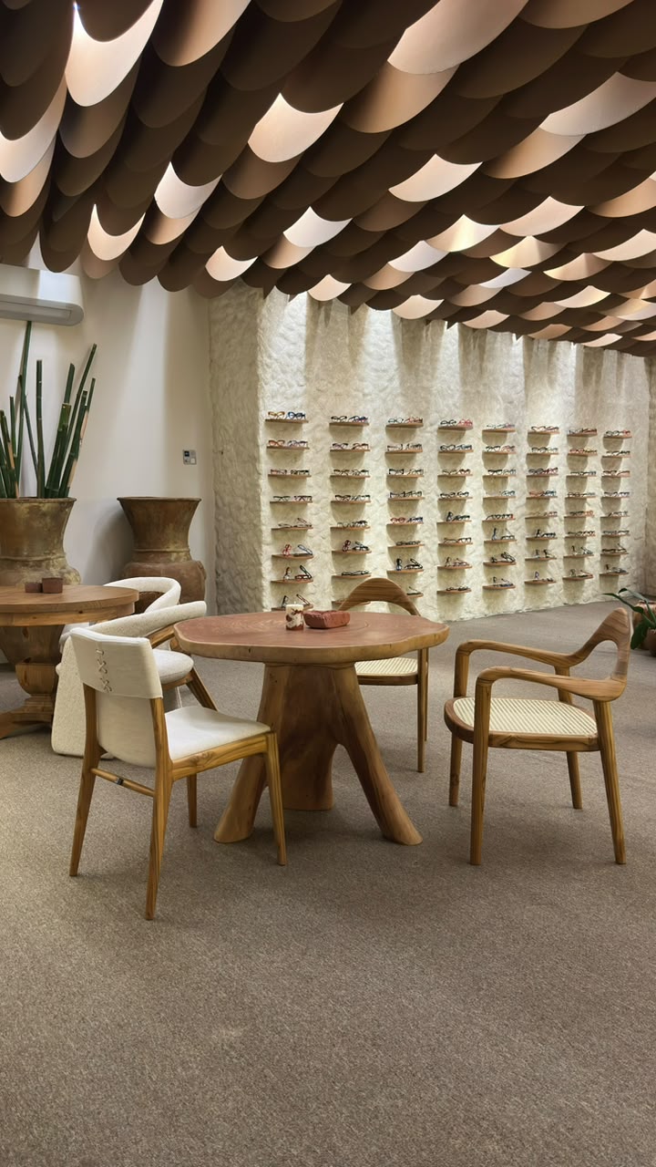 A modern dining or lounge area featuring a round wooden table with a unique trunk-like base, surrounded by wooden chairs with cream cushions, set in a stylish interior with a sculptural ceiling and a display wall of small wall-mounted objects.