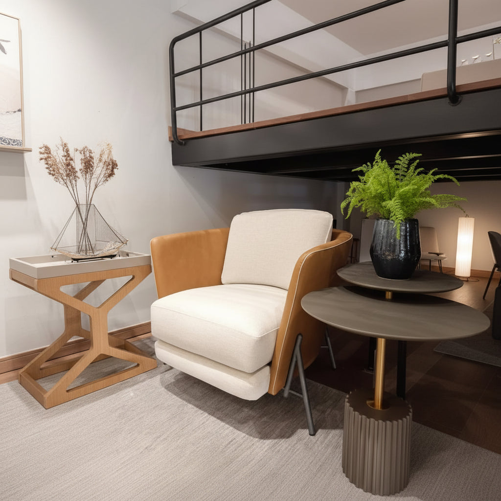 A modern accent chair with cream-colored fabric cushions and warm tan leather arms, styled in a contemporary loft setting next to a round side table.