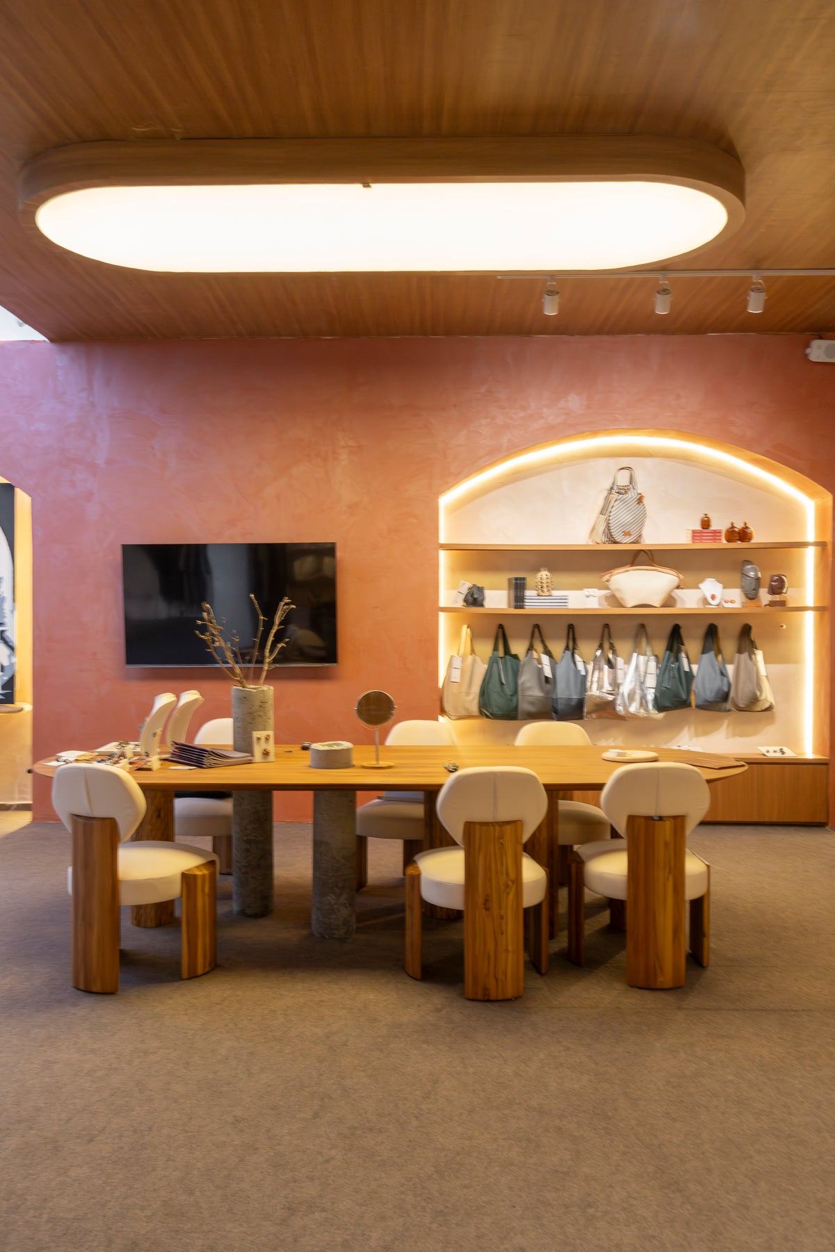 A modern dining area with a rectangular wooden table and six matching wooden chairs with cream upholstered seats, set against a terracotta-pink wall with built-in shelving and a TV.