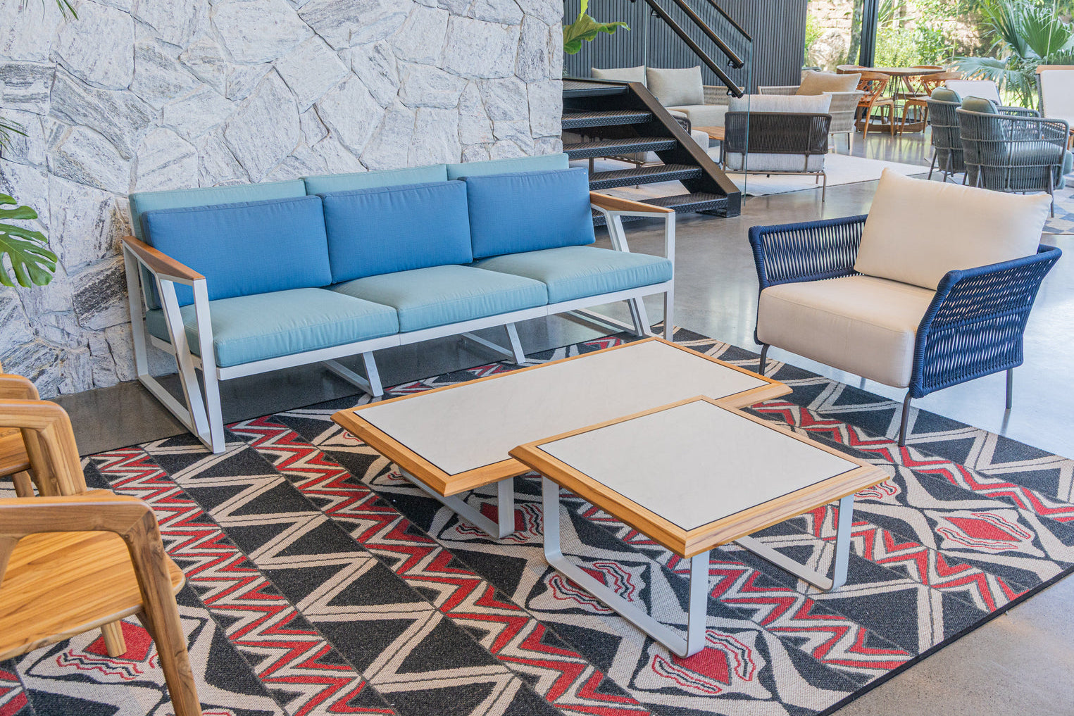 A modern outdoor lounge set displayed on a patterned rug in a showroom. The set includes a three-seat sofa with blue cushions and light wood arms, a matching armchair with a cream cushion and blue woven frame, and two nesting coffee tables with cream tops and light wood frames.