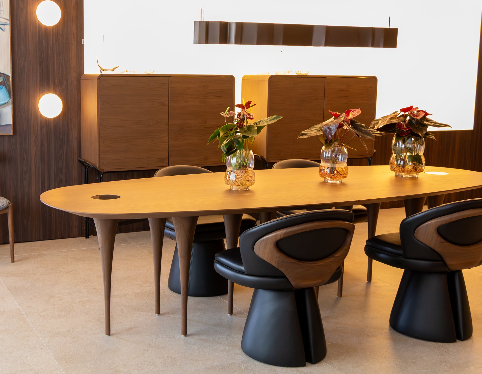 A modern dining room setup featuring a light wood oval dining table with sculptural black and wood chairs, decorated with three glass vases containing red and green plants, set against a dark wood cabinet and a white wall.