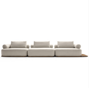 A modern modular three-seater sofa in a light beige fabric with matching back cushions and a small wooden accent board under the right side, shown against a plain white background.