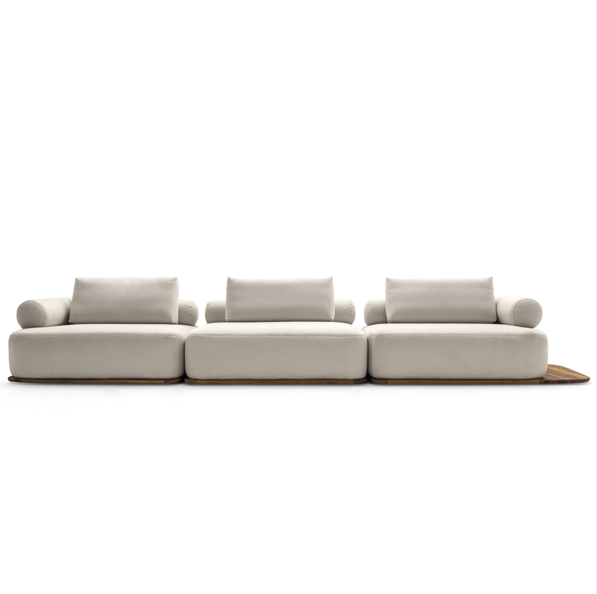 A modern modular three-seater sofa in a light beige fabric with matching back cushions and a small wooden accent board under the right side, shown against a plain white background.