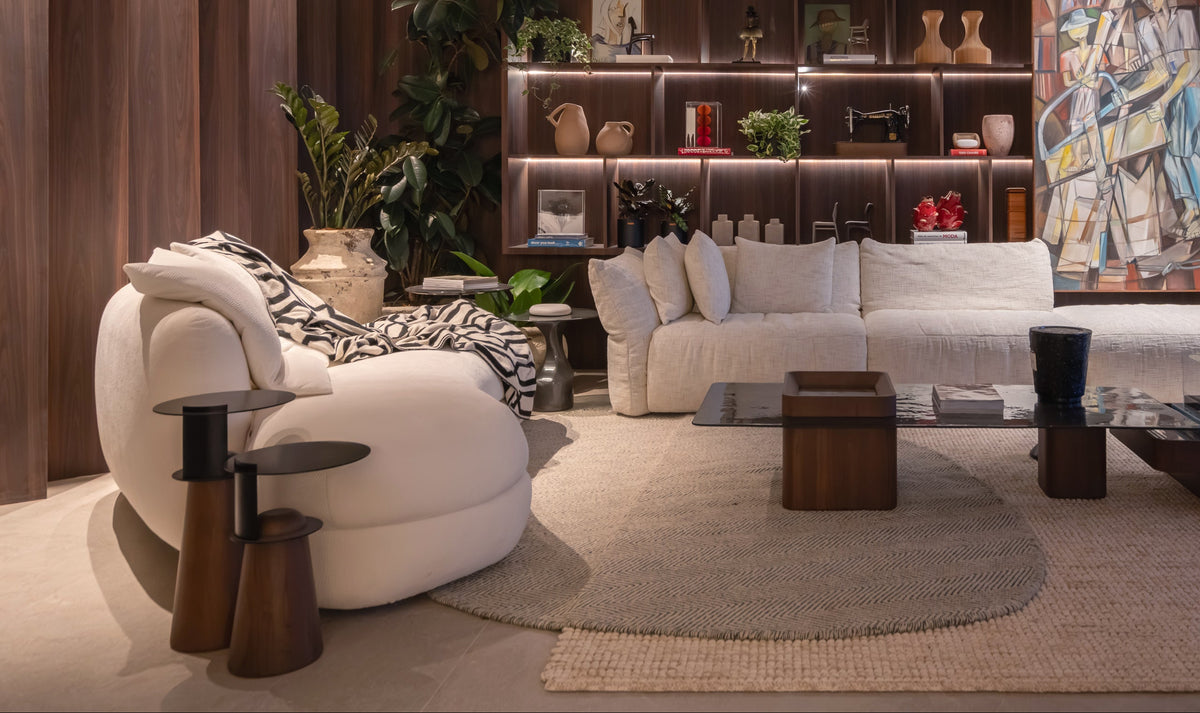 A modern, stylishly decorated living room featuring a white curved lounge chair, a white sectional sofa, dark wood shelving, and a large colorful abstract painting on the wall.
